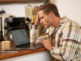 Image of adult man looking complicated at laptop screen, staring puzzled at his monitor, doing difficult task online, sitting in cafe, drinking coffee and working
